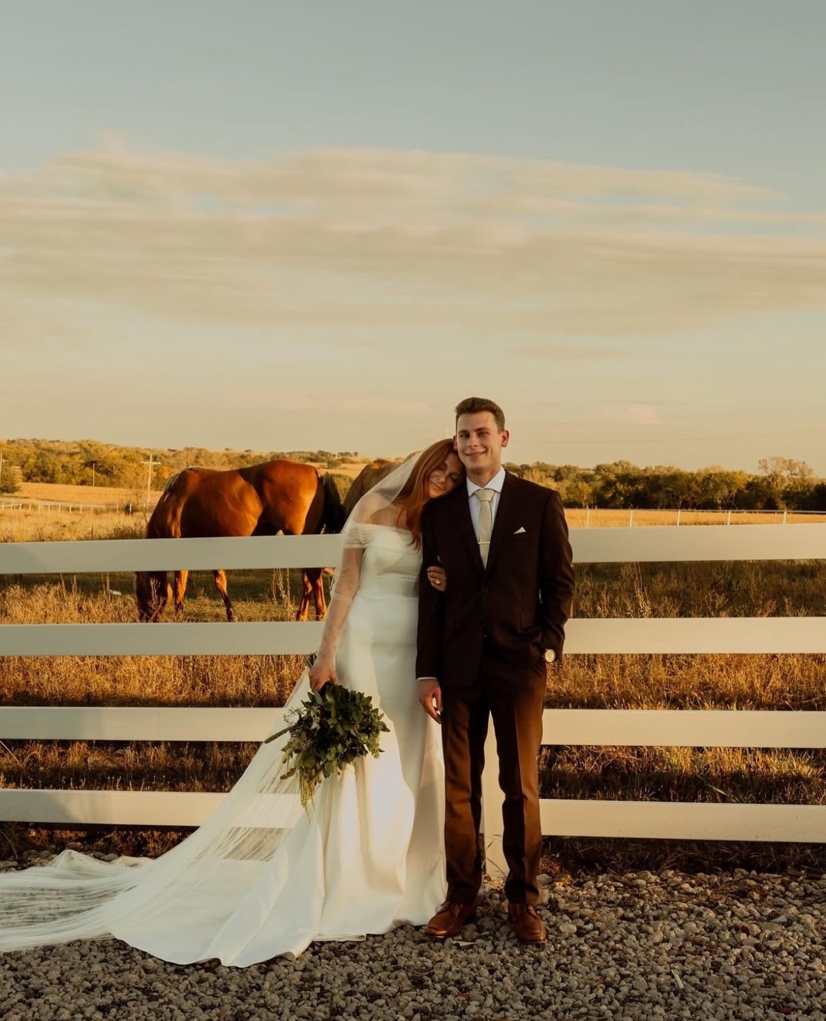 Bride and groom pose by white fence at outdoor Nebraska wedding venue with horses and pasture backdrop