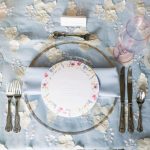 Elegant place setting with floral-rimmed plate on glass charger atop blue floral tablecloth with vintage silverware
