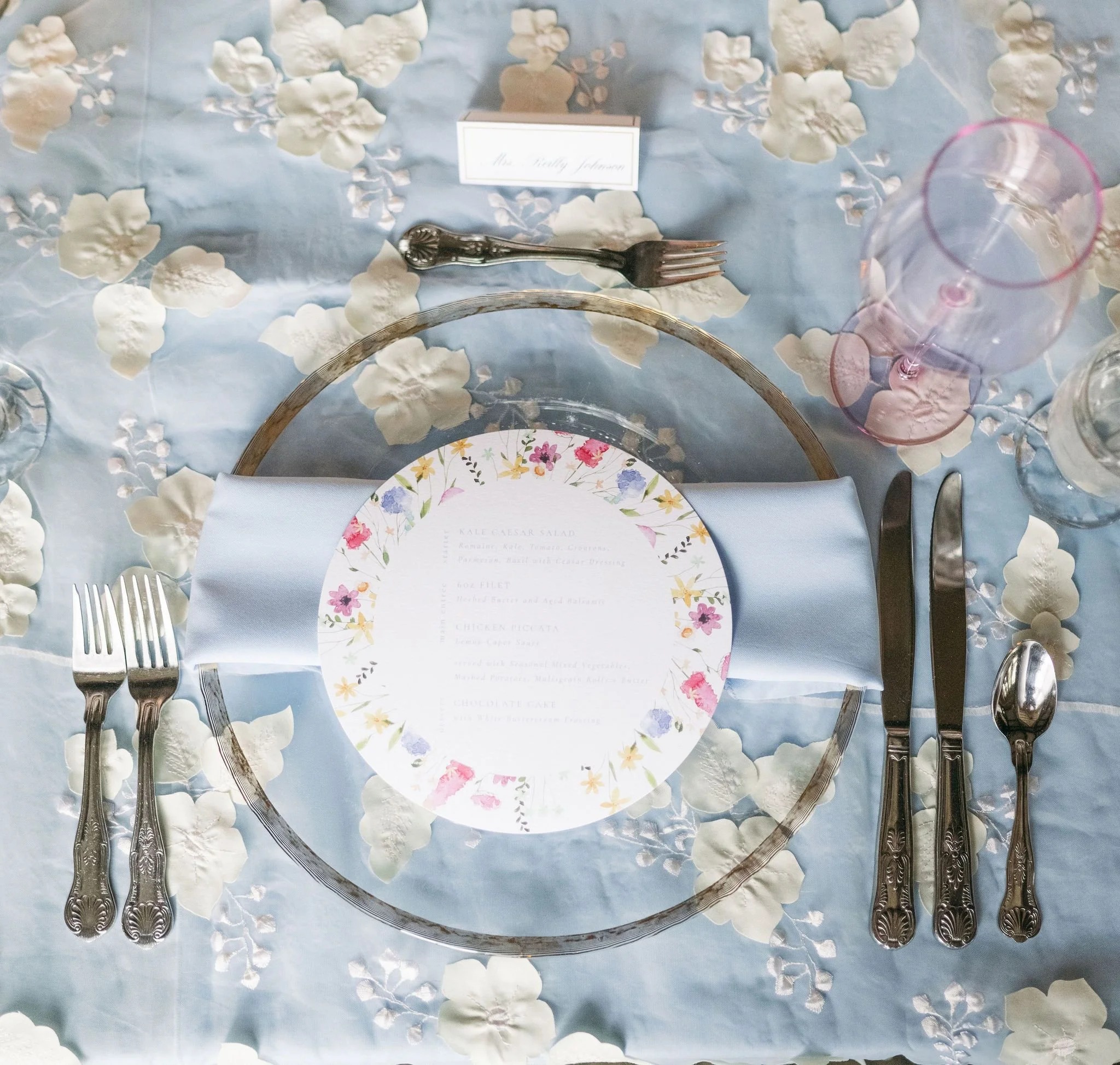 Elegant place setting with floral-rimmed plate on glass charger atop blue floral tablecloth with vintage silverware