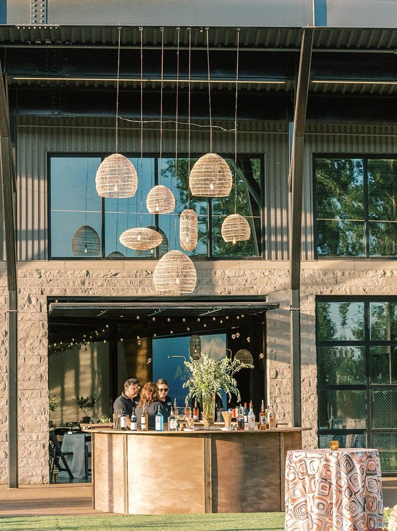 Outdoor bar setup with woven pendant lights, wooden bar, and patterned cocktail table linens