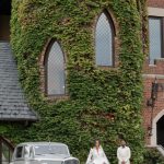 Newlywed couple standing beside classic vintage car in front of ivy-covered Gothic-style building