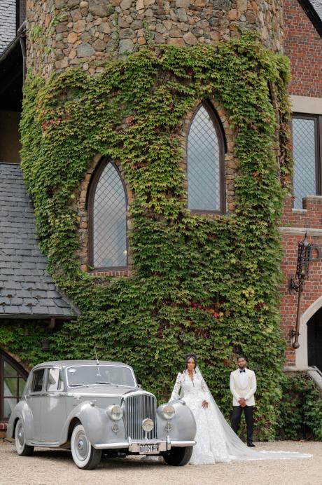 Newlywed couple standing beside classic vintage car in front of ivy-covered Gothic-style building