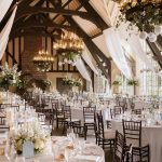 Rustic barn wedding reception hall with exposed beam vaulted ceiling, elegant chandeliers with greenery, flowing white drapes, and round tables set with white linens