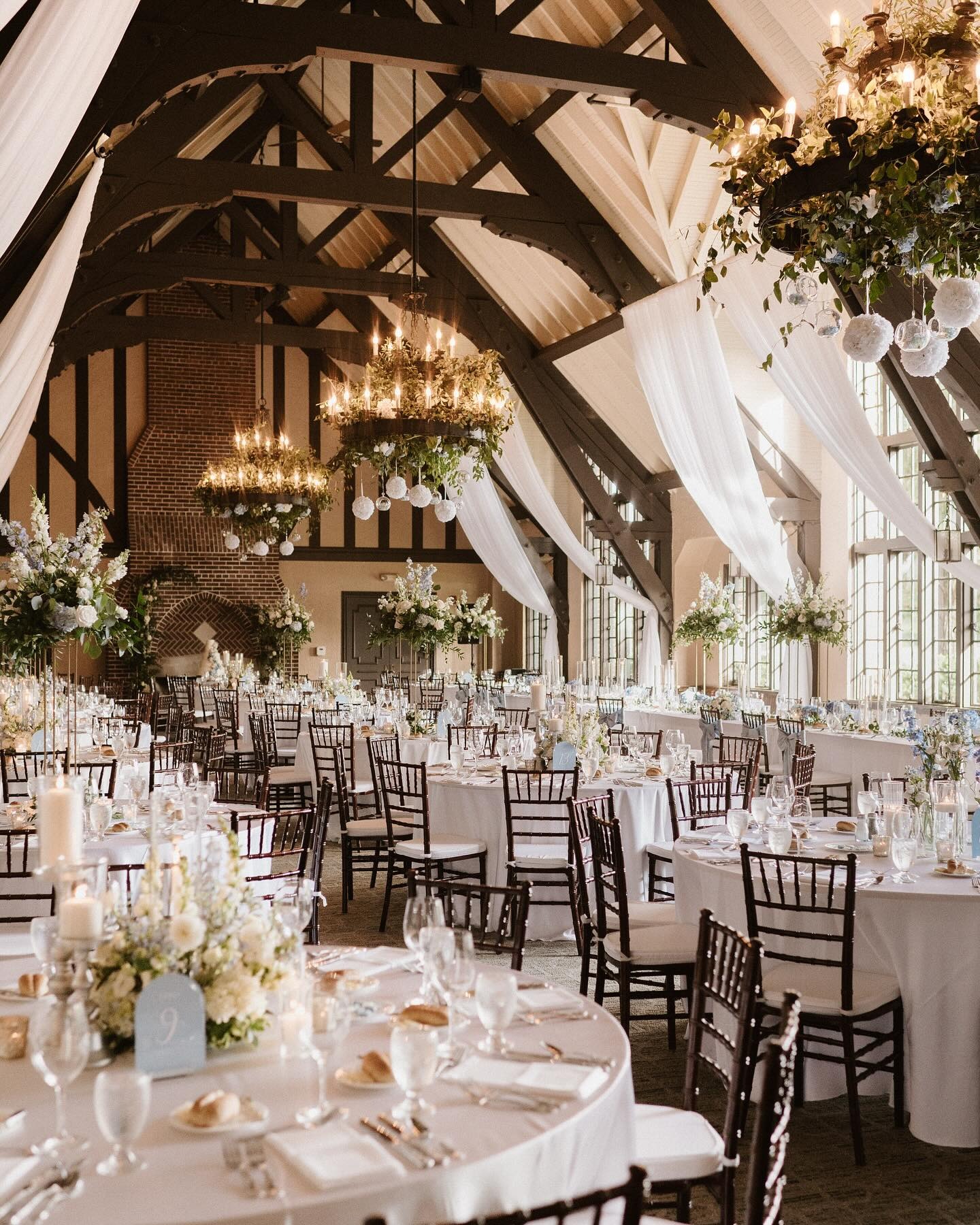 Rustic barn wedding reception hall with exposed beam vaulted ceiling, elegant chandeliers with greenery, flowing white drapes, and round tables set with white linens