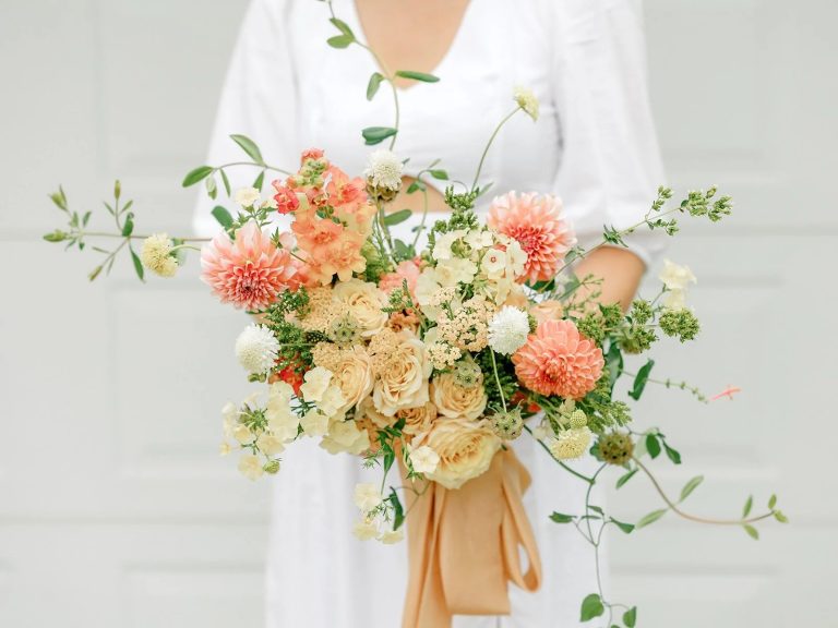 Wedding bouquet with coral dahlias, cream roses, and greenery tied with gold silk ribbon
