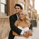 Bride with wavy blonde hair and natural makeup embraces groom in black suit at urban brick venue