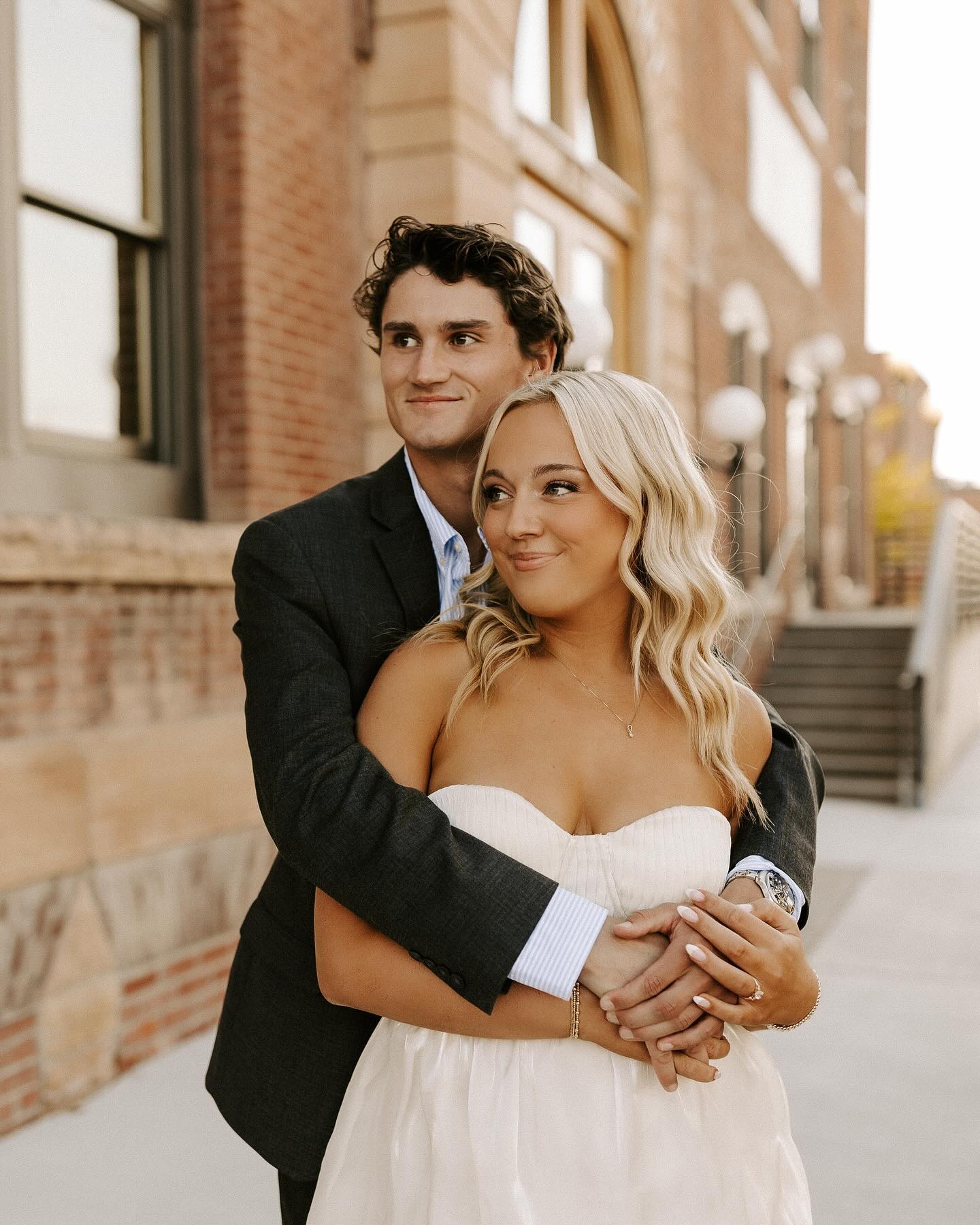 Bride with wavy blonde hair and natural makeup embraces groom in black suit at urban brick venue