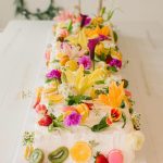 Three-tier wedding cake with white frosting decorated with colorful edible flowers, fresh fruit slices, and macarons