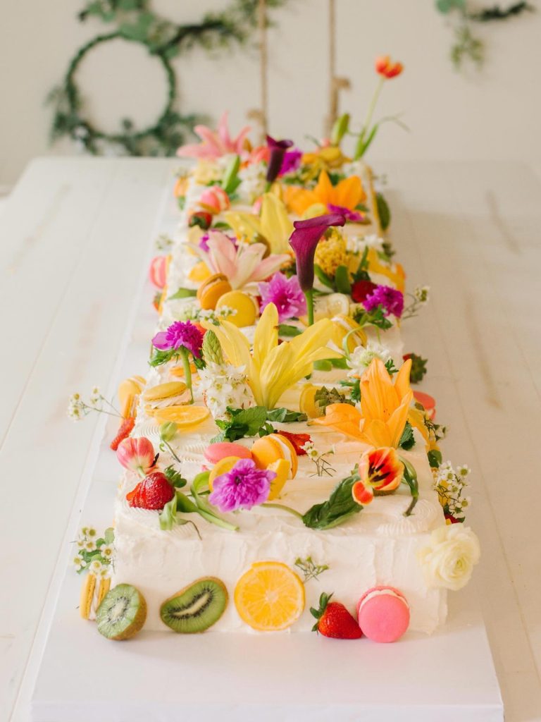 Three-tier wedding cake with white frosting decorated with colorful edible flowers, fresh fruit slices, and macarons