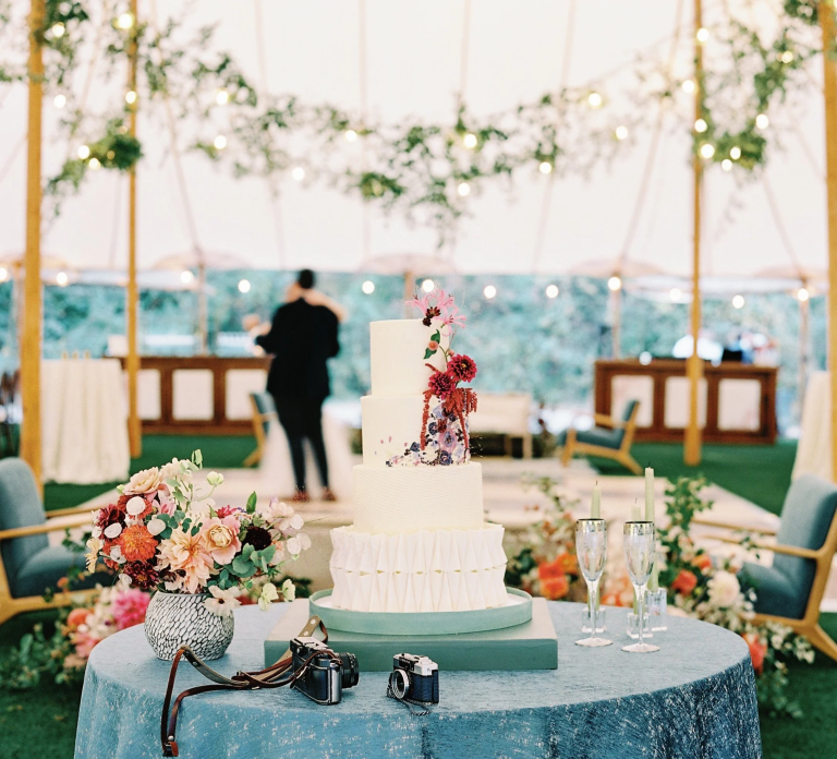 Three-tier wedding cake with vertical pleating, floral cascade decoration, and turquoise accents displayed at tent reception