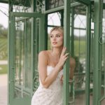Bride in embroidered wedding dress posing beside sage green glass door in ornate greenhouse
