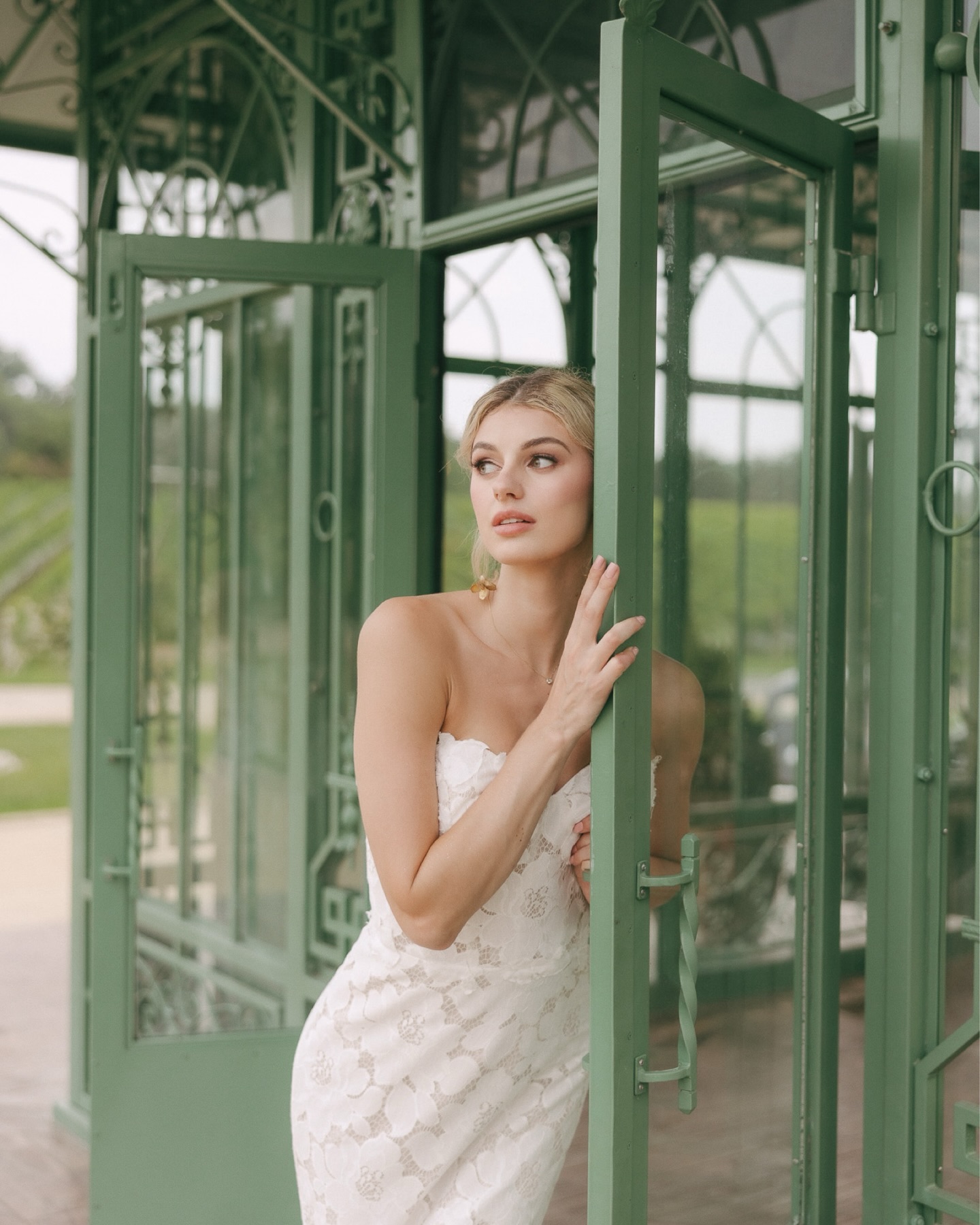 Bride in embroidered wedding dress posing beside sage green glass door in ornate greenhouse