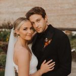 Newlywed couple embracing outdoors, bride in sleek spaghetti strap gown with veil, groom in black suit