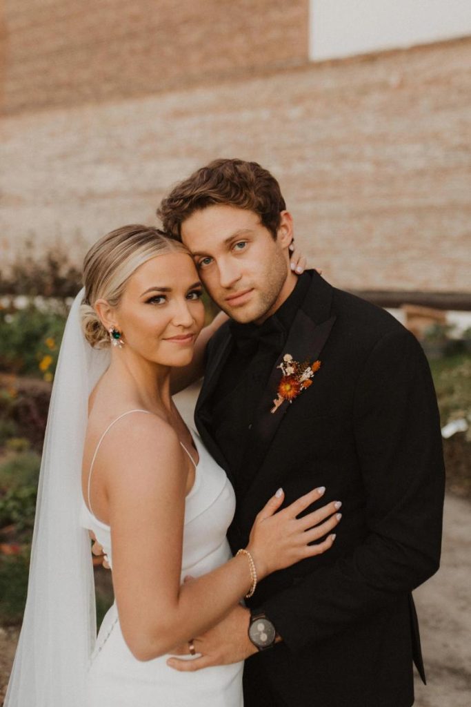 Newlywed couple embracing outdoors, bride in sleek spaghetti strap gown with veil, groom in black suit