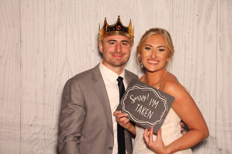 Wedding couple posing in photo booth with props and "Sorry I'm Taken" sign against white wood backdrop