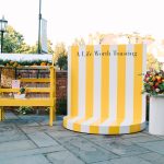 Yellow and white striped circular bar display reading "A Life Worth Toasting" with yellow beverage station and colorful flower arrangement