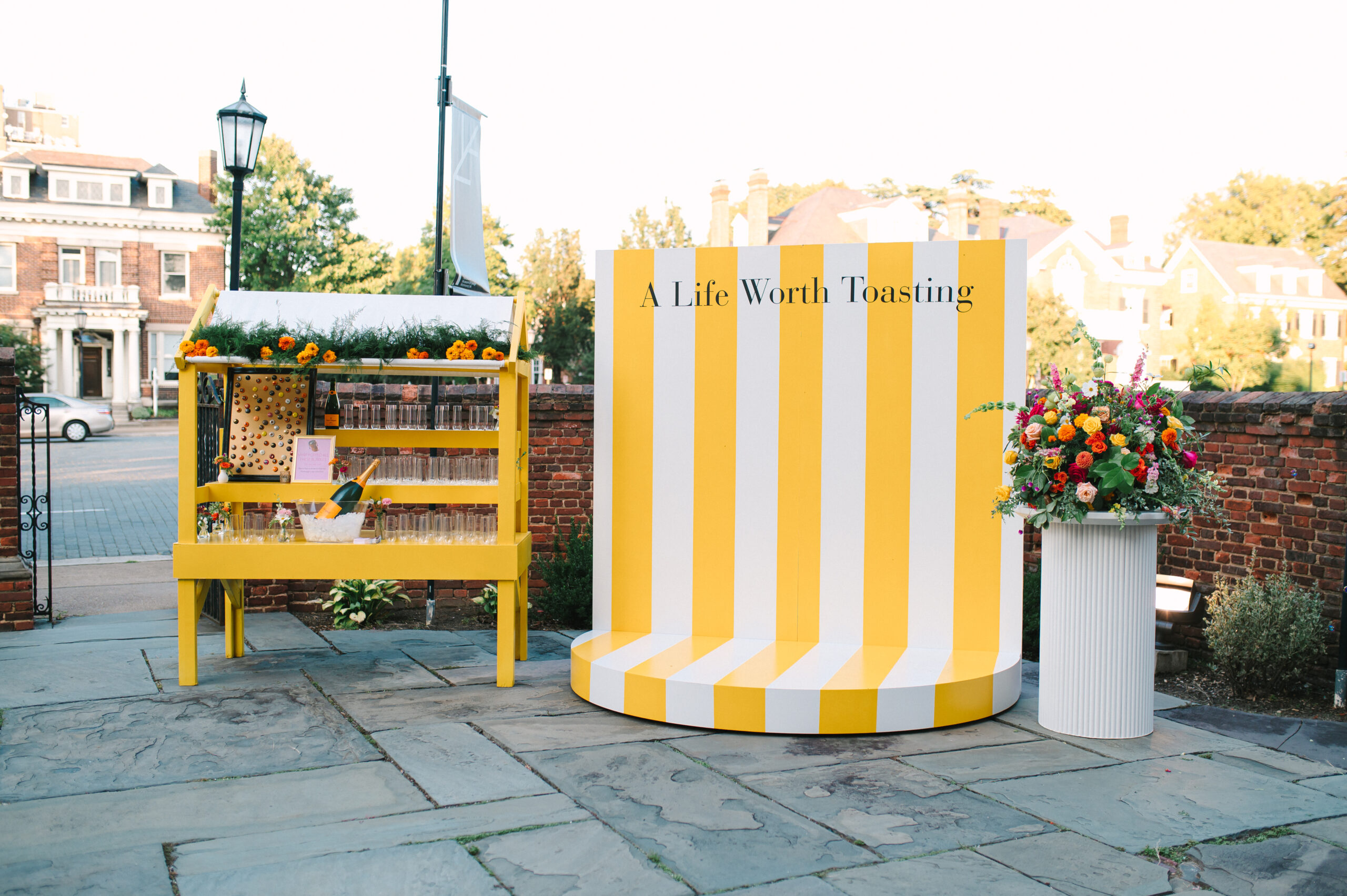 Yellow and white striped circular bar display reading "A Life Worth Toasting" with yellow beverage station and colorful flower arrangement