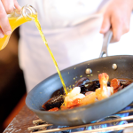 Chef pouring olive oil into sauté pan with seafood over stovetop burner