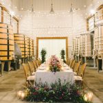 Winery barrel room dinner setup with string lights, long table, cane-back chairs, and lush purple floral arrangements