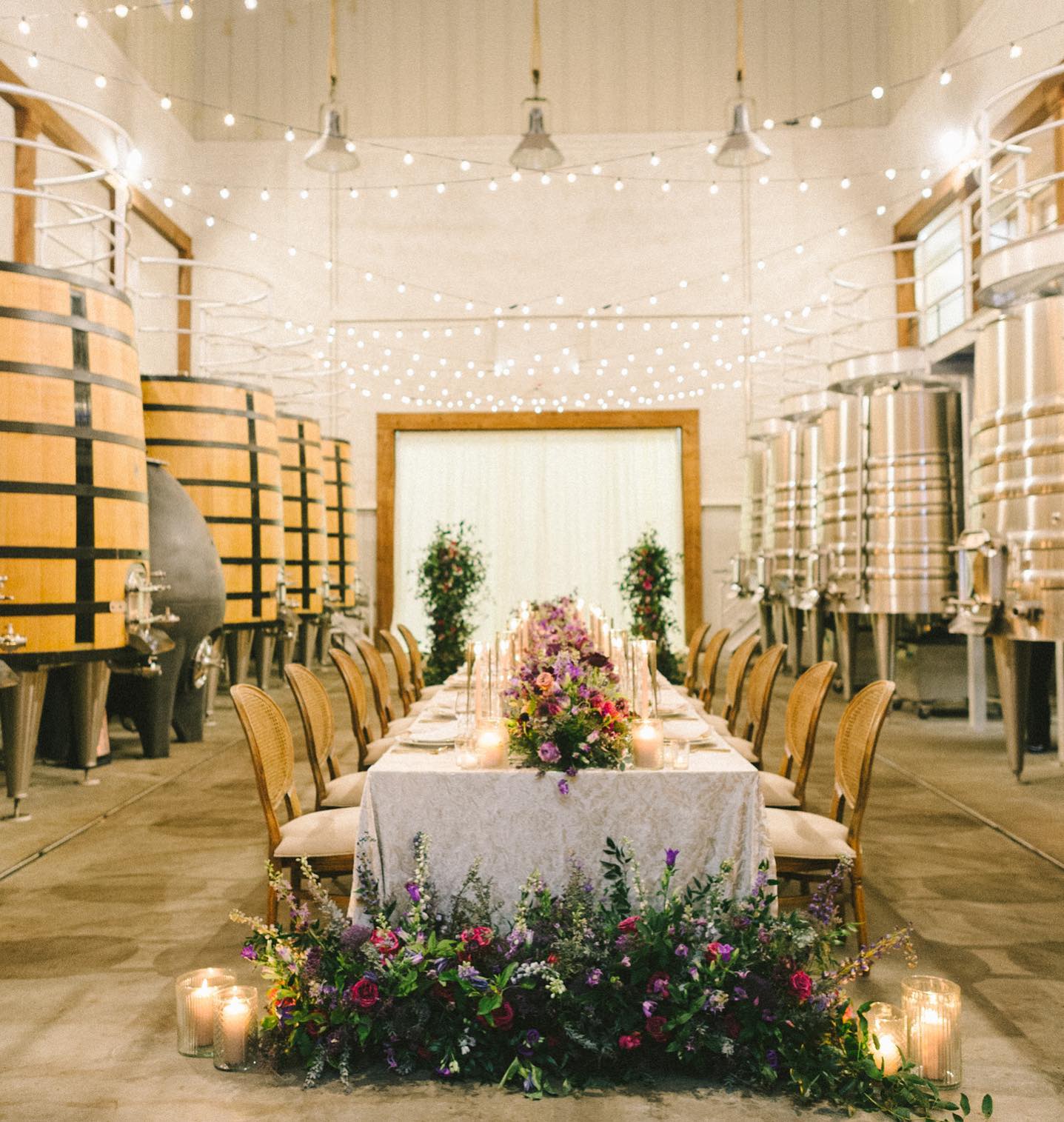 Winery barrel room dinner setup with string lights, long table, cane-back chairs, and lush purple floral arrangements