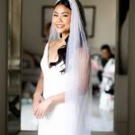 Smiling bride in white wedding dress and pearl-adorned veil with polished makeup and styled dark hair
