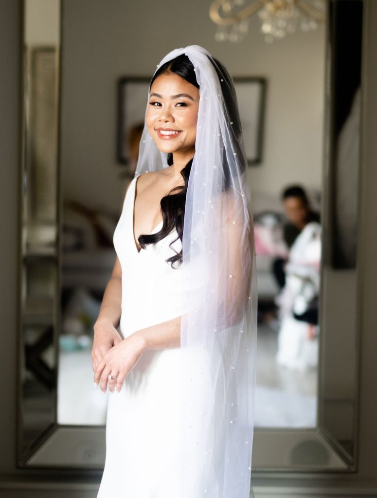Smiling bride in white wedding dress and pearl-adorned veil with polished makeup and styled dark hair