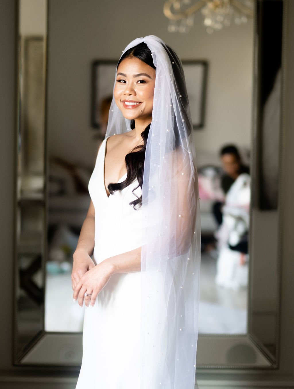 Smiling bride in white wedding dress and pearl-adorned veil with polished makeup and styled dark hair
