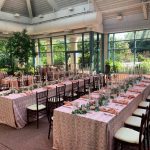 Long banquet tables with pink patterned linens and greenery centerpieces arranged in glass conservatory with garden views