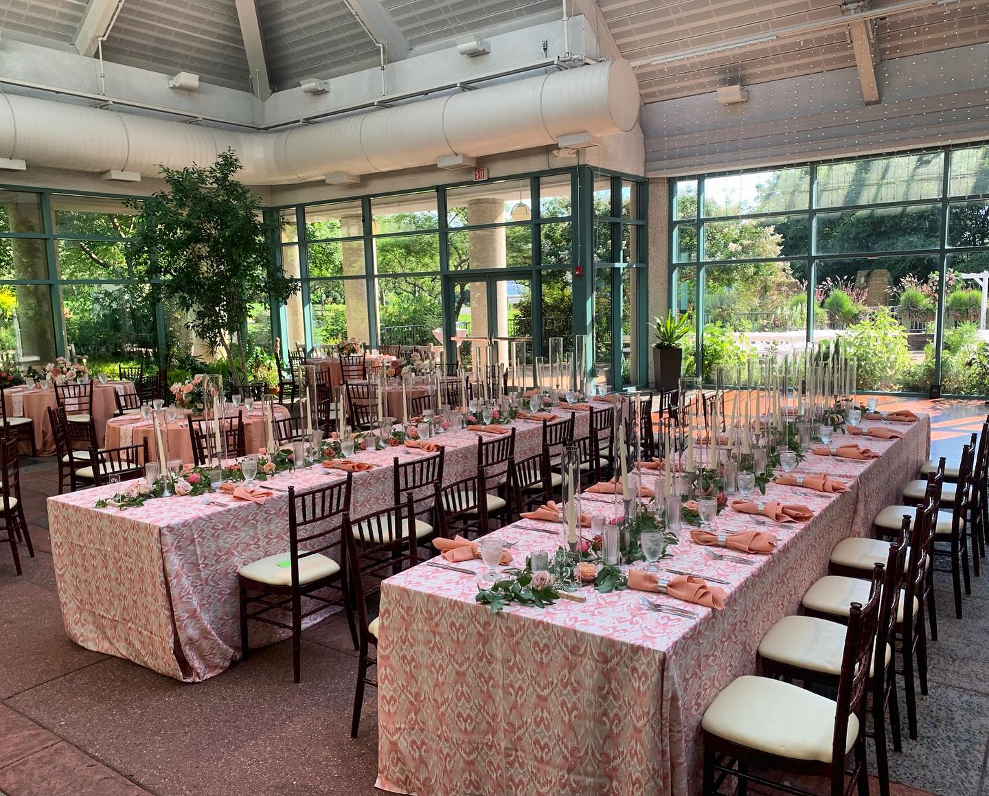 Long banquet tables with pink patterned linens and greenery centerpieces arranged in glass conservatory with garden views