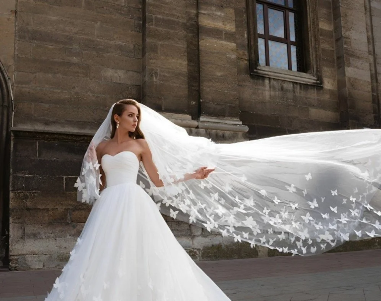 Bride in strapless ball gown with dramatic butterfly-embellished cathedral veil billowing in wind