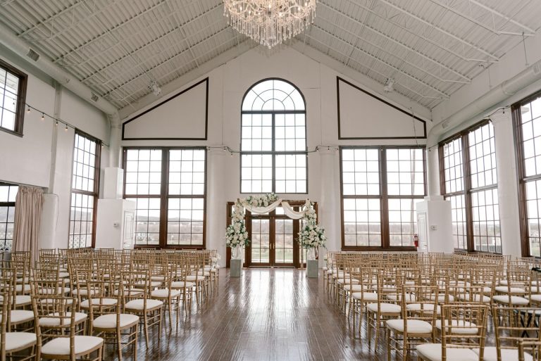 Industrial wedding ceremony space with arched windows, crystal chandelier, and gold chiavari chairs