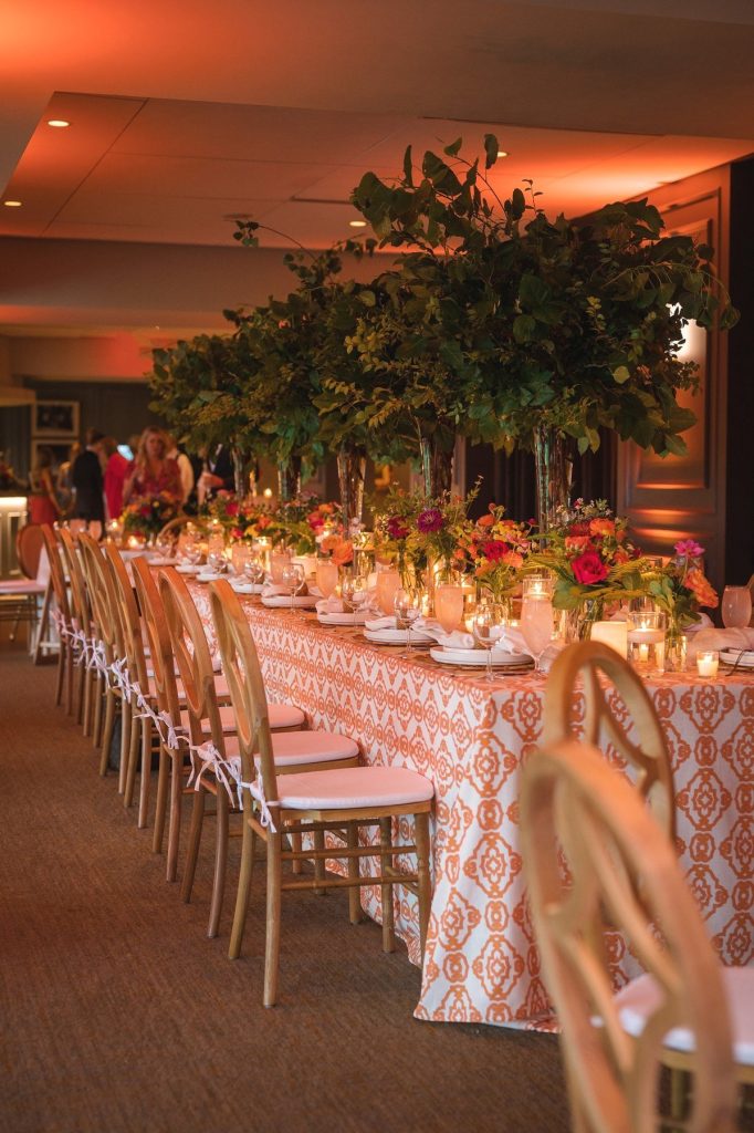 Long reception table with orange patterned linens, gold chiavari chairs, tall green centerpieces, and candlelight under warm amber uplighting