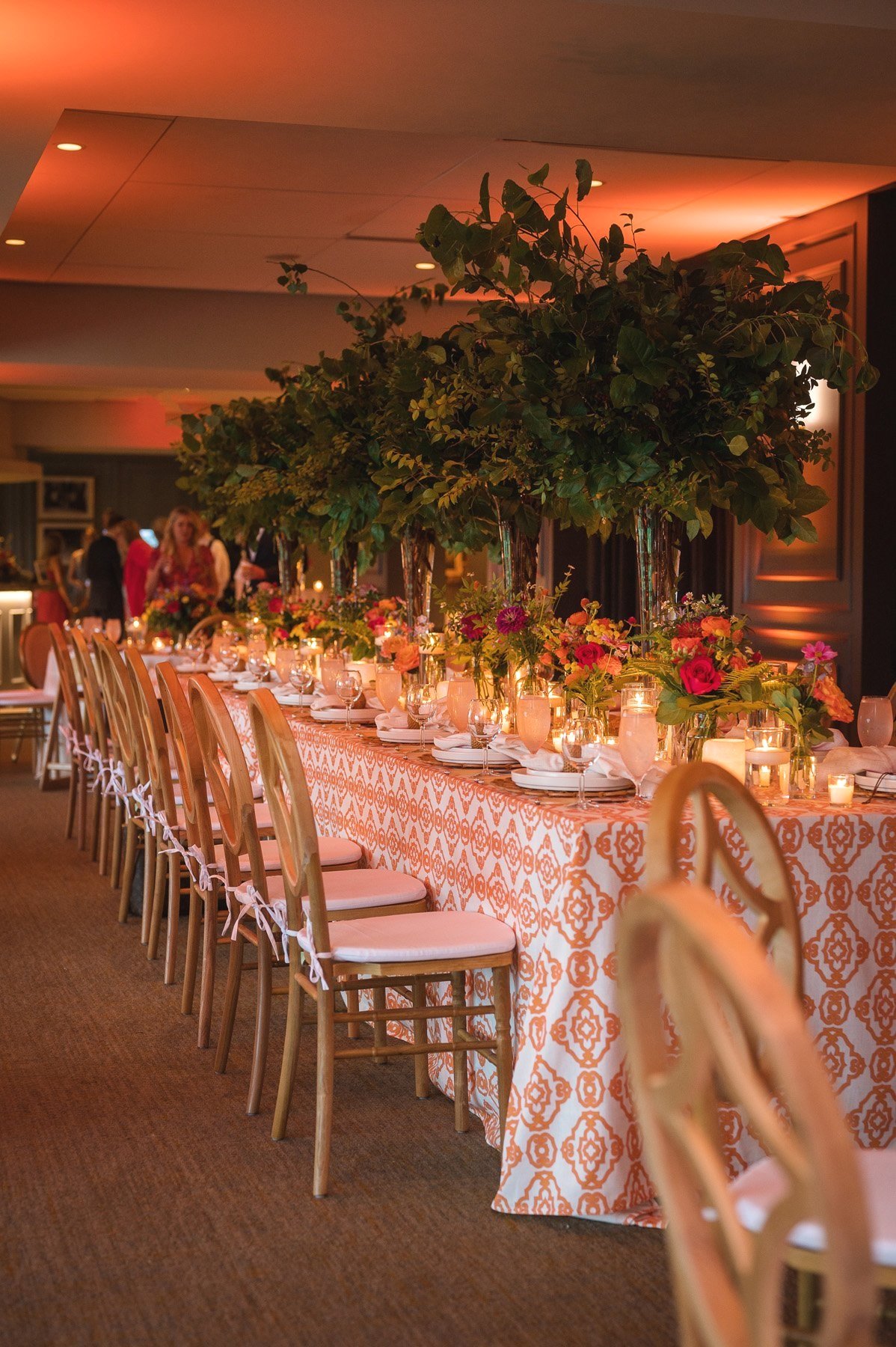 Long reception table with orange patterned linens, gold chiavari chairs, tall green centerpieces, and candlelight under warm amber uplighting