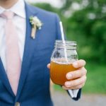 Groom in blue suit with boutonniere holding mason jar cocktail with straw at outdoor wedding