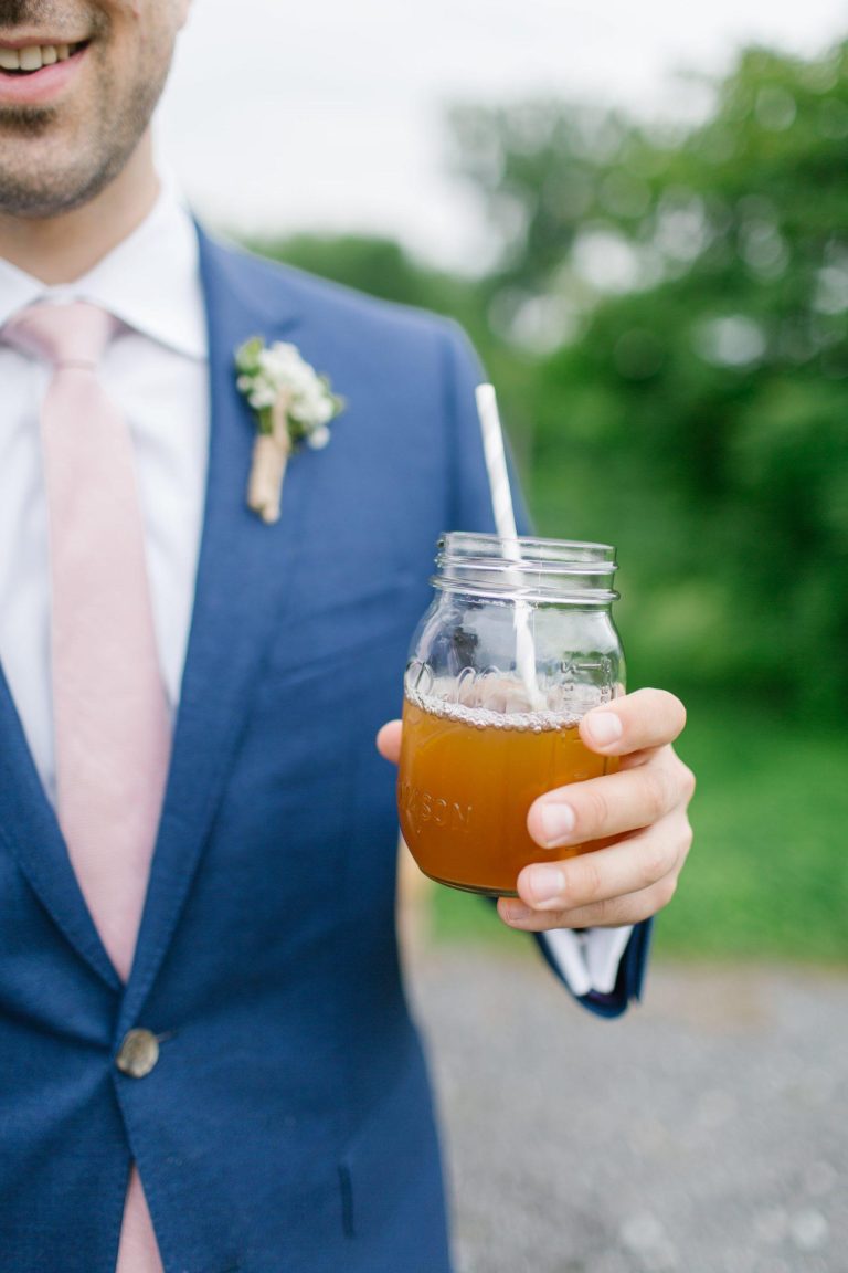 Groom in blue suit with boutonniere holding mason jar cocktail with straw at outdoor wedding