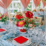 Elegant wedding table setting with red and coral floral centerpieces, blue linens, and coral draped tent