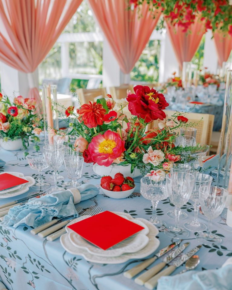 Elegant wedding table setting with red and coral floral centerpieces, blue linens, and coral draped tent