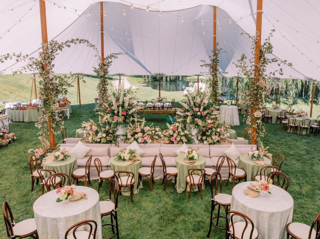 Elegant tented wedding reception with wooden bistro chairs, ivory linens, pink floral centerpieces, lounge seating, and lush greenery garlands
