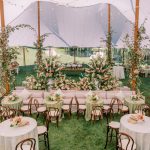 Elegant tented wedding reception with wooden bistro chairs, ivory linens, pink floral centerpieces, lounge seating, and lush greenery garlands