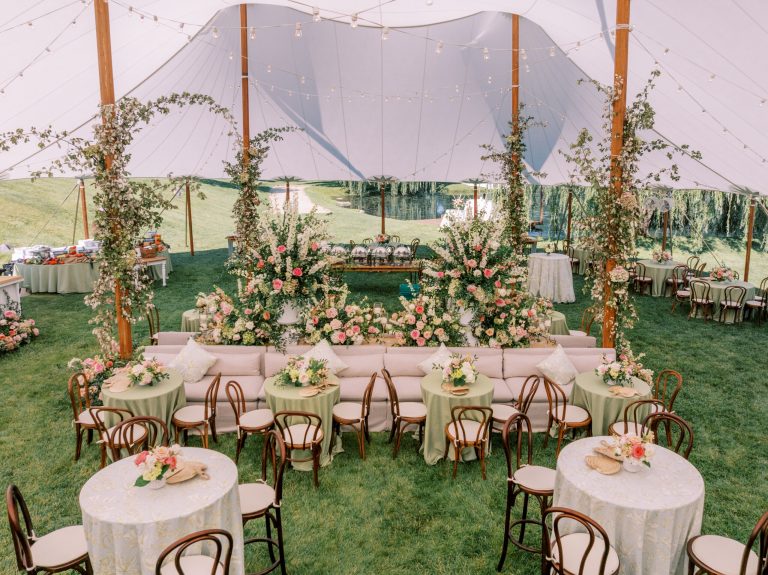 Elegant tented wedding reception with wooden bistro chairs, ivory linens, pink floral centerpieces, lounge seating, and lush greenery garlands