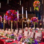 Dramatic banquet table with tall gold candelabras, vibrant red and purple floral arrangements, and red sequin tablecloth