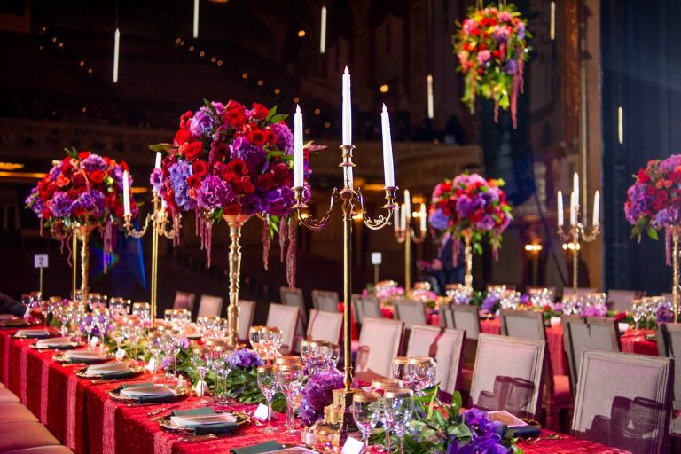 Dramatic banquet table with tall gold candelabras, vibrant red and purple floral arrangements, and red sequin tablecloth