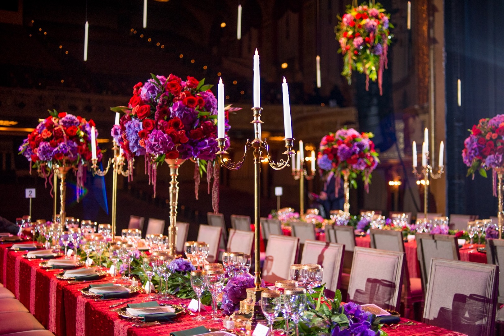 Dramatic banquet table with tall gold candelabras, vibrant red and purple floral arrangements, and red sequin tablecloth