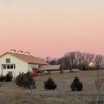 White barn wedding venue with three cupolas set on rural farmland at sunset with evergreen trees and bare winter trees in background