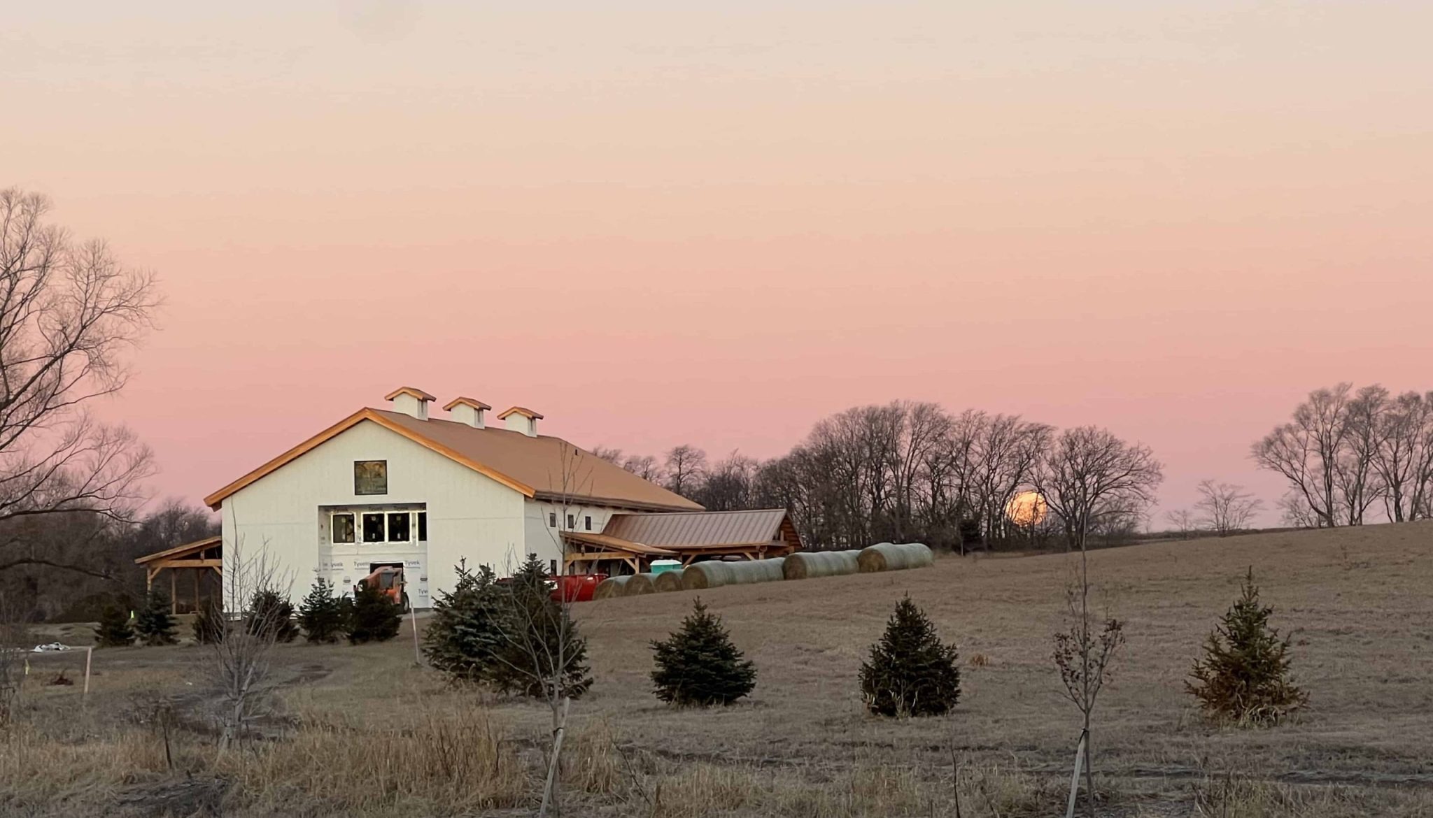 White barn wedding venue with three cupolas set on rural farmland at sunset with evergreen trees and bare winter trees in background