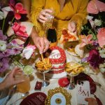 Overhead view of celebratory table spread with champagne, pizza, fries, red cake, and tropical flowers surrounding guest in yellow fringed outfit
