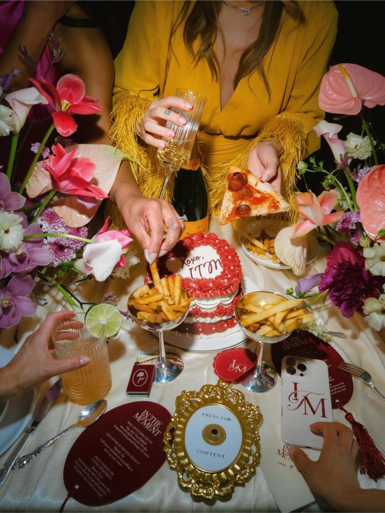 Overhead view of celebratory table spread with champagne, pizza, fries, red cake, and tropical flowers surrounding guest in yellow fringed outfit