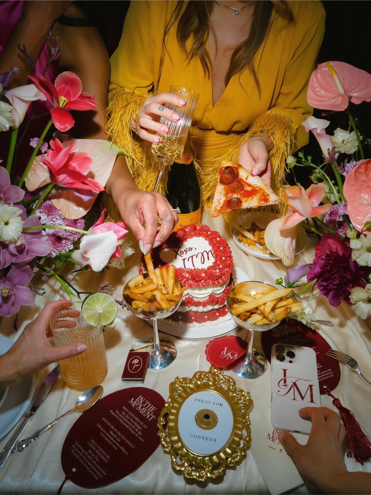 Overhead view of celebratory table spread with champagne, pizza, fries, red cake, and tropical flowers surrounding guest in yellow fringed outfit