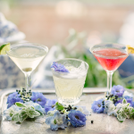Three cocktails on silver tray with blue and white flowers and citrus garnishes