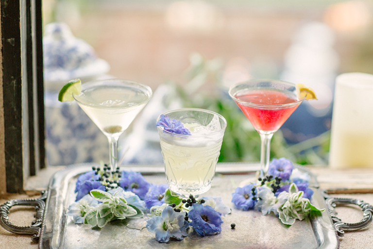 Three cocktails on silver tray with blue and white flowers and citrus garnishes